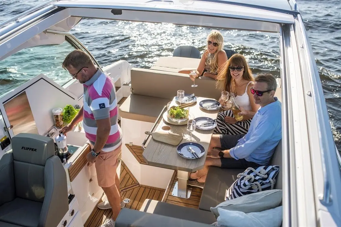 A group of four friends enjoys drinks at a stylish boat kitchen, with a man preparing food and a table set for dining.